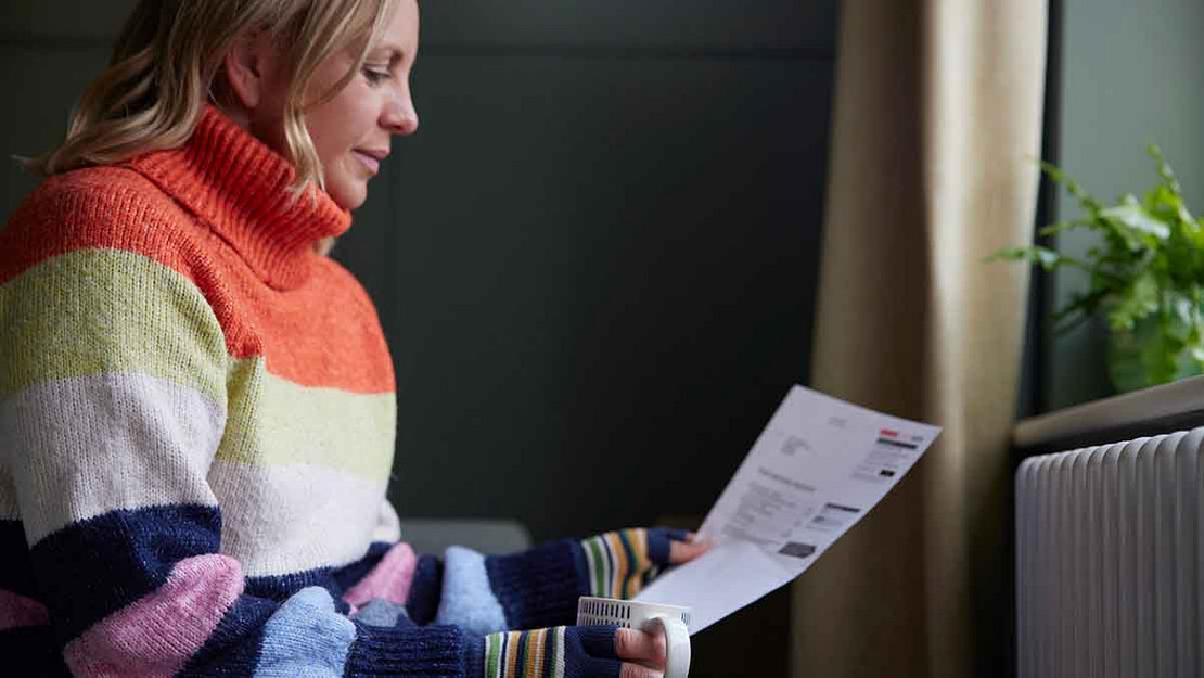 Frau im dicken Pullover sitzt vor der Heizung und hält ein Schreiben in der Hand. 